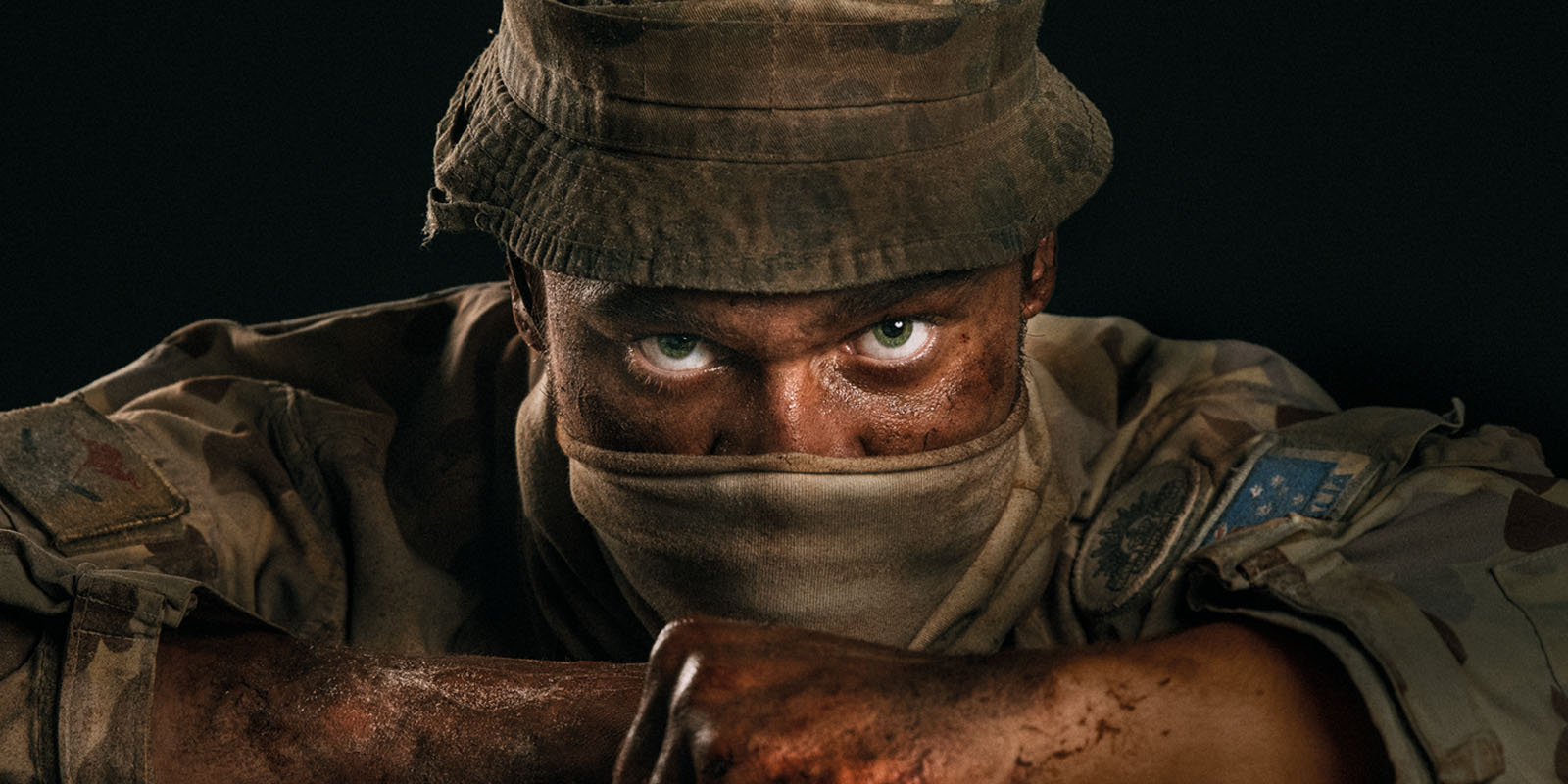 Headshot of Australian soldier in uniform, dirty and sweaty, with mouth covered and intense green eyes
