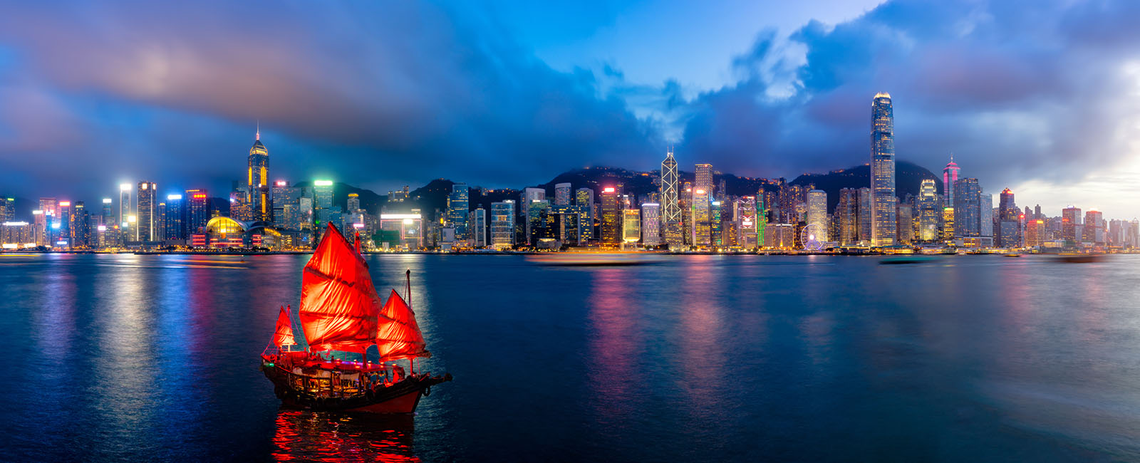 A traditional junk boat with red sails floats in Hong Kong harbour at night. City Skyline in background/  