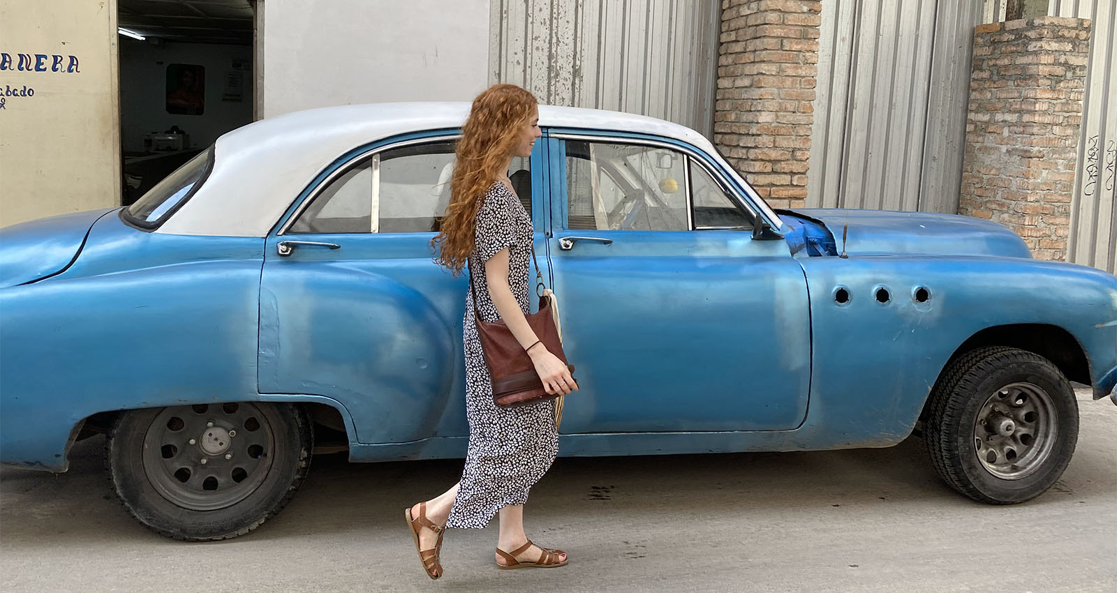 A lady in a long dress with long red hair walks past a blue and white vintage car 