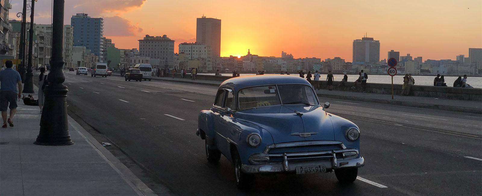 A vintage car on the road with the sun setting behind the city skyline