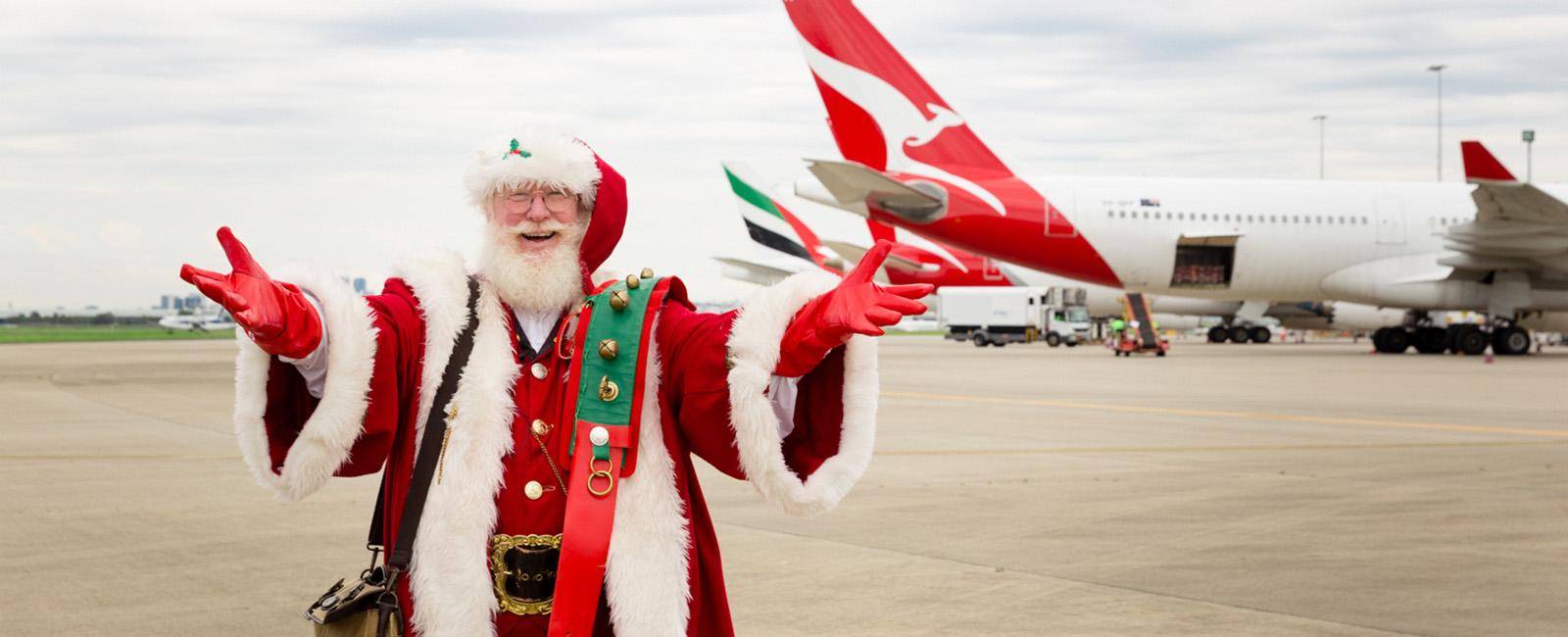 Santa Claus standing with arms wide open and smiling face in front of two airplanes at Brisbane airport