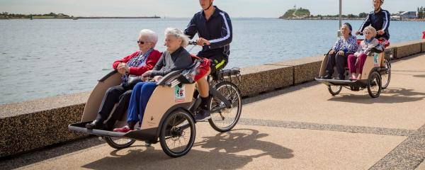 two cyclists with elderly passengers cycling along a path near water
