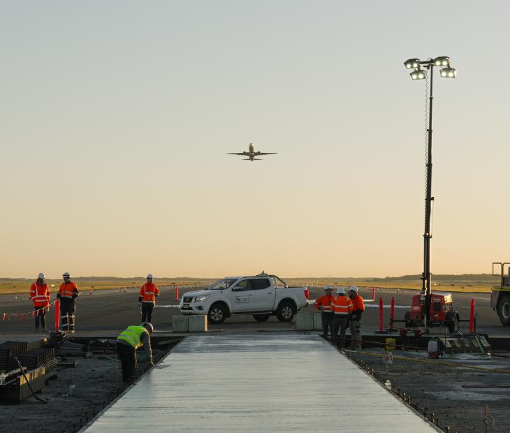 Construction workers pouring concrete on the Legacy Runway with a plane flying overhead Construction workers pouring concrete on the Legacy Runway with a plane flying overhead