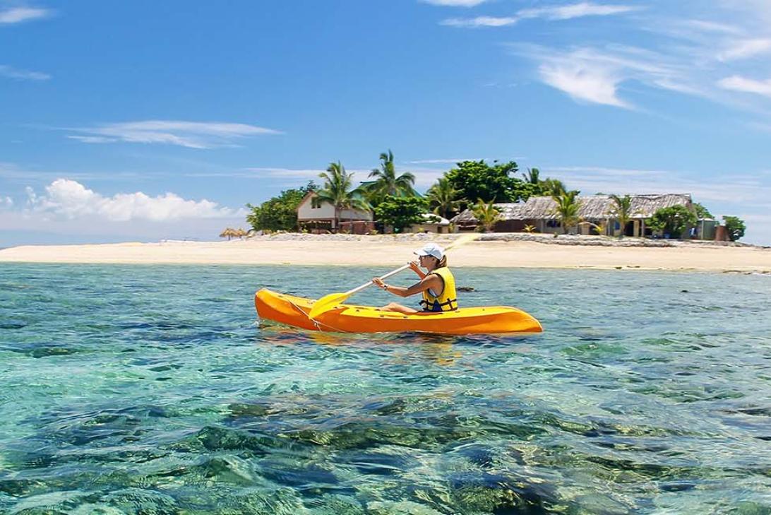 A person in a yellow kayak on sparkly blue, clear waters. A small tropical island with a beach and palm trees is in the background. 